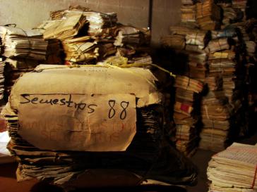 Unmengen aufgestapelter Dokumente, einzelner Blätter und Papierfetzen füllen die Räume des Archiv. Hier im Bild ein Verweis mit der Beschriftung ''Manuskripte 1988. Entführungen.'' Aufgetürmte Unterlagen im Archiv